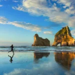 A person running along a wet sandy beach at low tide, with their reflection visible on the surface. In the background, large rock formations rise from the ocean—one featuring a natural arch—under a blue sky with scattered clouds lit by warm sunlight.